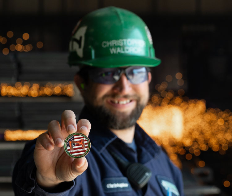 Smiling male Vulcraft employee with dark beard wearing green hard hat with award coin.