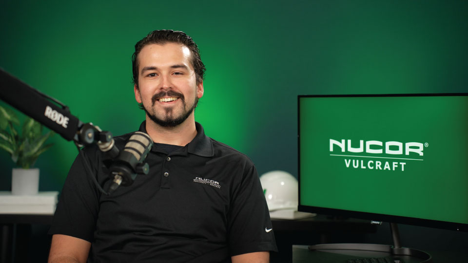 Vulcraft Verco's Doug Eicher in black Nucor polo shirt smiling with a mic in front of him. Green background & Nucor Vulcraft logo on a monitor behind him to the right. Cover image for Floor Wizard Design Tool video.