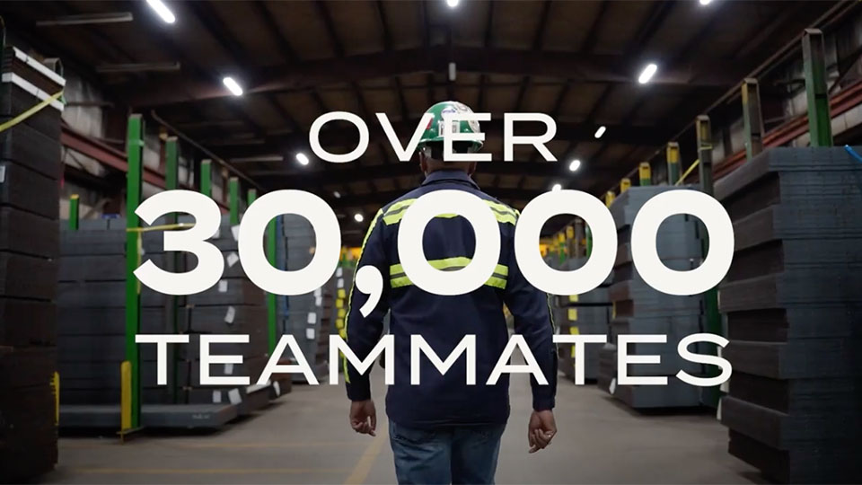 Nucor promo video cover image showing Nucor teammate wearing a blue safety uniform and green safety hat walking between rows of steel in a plant.