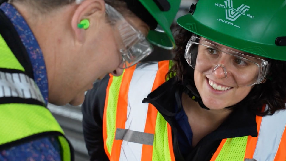Closeup image of a smiling female Verco teammate with shoulder-length dark hair in green hardhat, safety glasses, and reflective safety vest on the right side of the frame, looking at a male teammate on the left side of the frame. He's faced away from the camera, but is wearing safety glasses, earplugs, and a reflective safety vest.